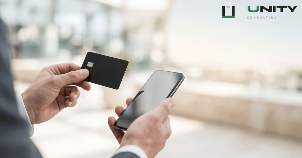 A close-up of hands holding a plain payment card and a smartphone, with a bright, blurred background.