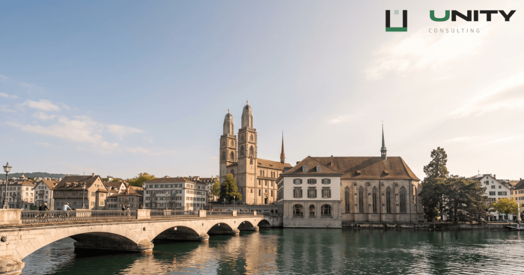 A panoramic view of Zurich, Switzerland, showing the Limmat River, historic city buildings, and church spires under a bright, clear sky.