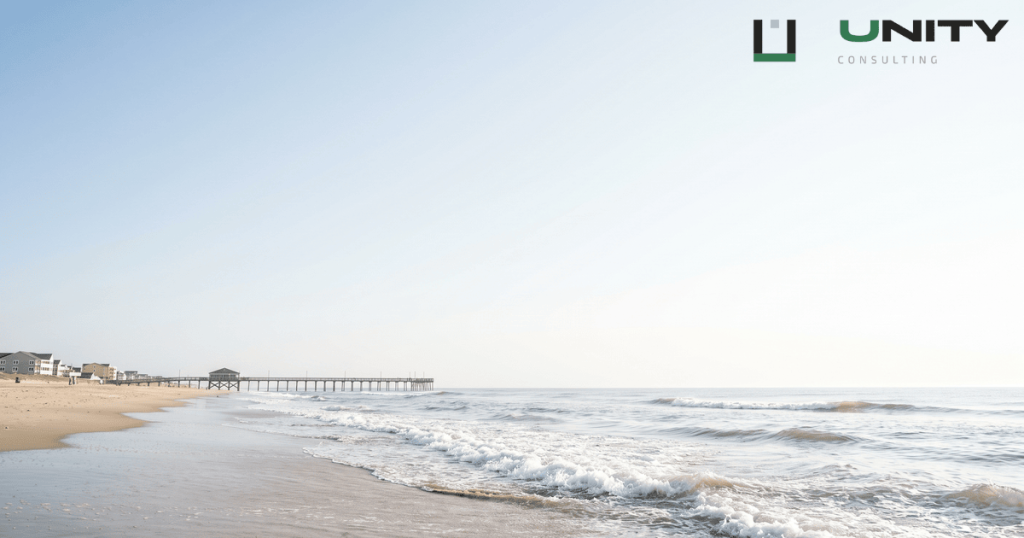 Wide shot of Rehoboth Beach, Delaware, with gentle waves on the sand and a distant pier under a bright, open sky.