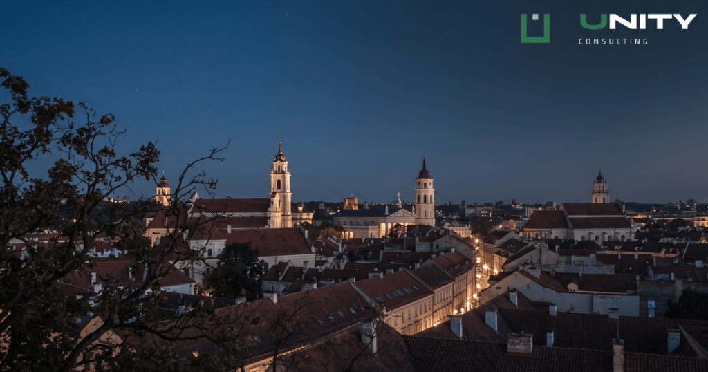 Twilight view over the red-tiled roofs and baroque spires of Vilnius Old Town with warm lights glowing.
