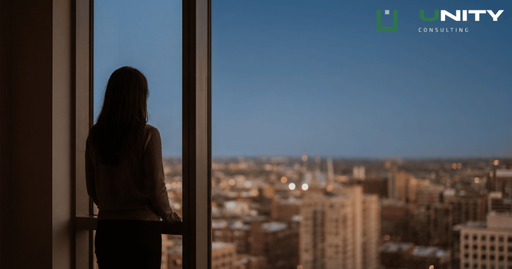 A person's silhouette stands at a window in a dim room, looking out at a softly lit city skyline at blue hour.