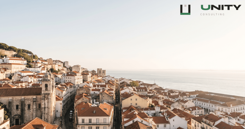 A wide, sunlit view of Lisbon's historic Alfama district and the Tagus River under a clear sky.