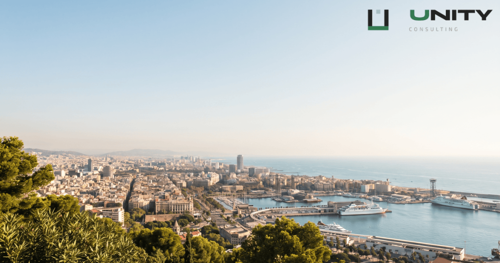 A wide, sun-drenched panoramic view of Barcelona's coastline and city from Montjuïc, with a bright sky overhead.