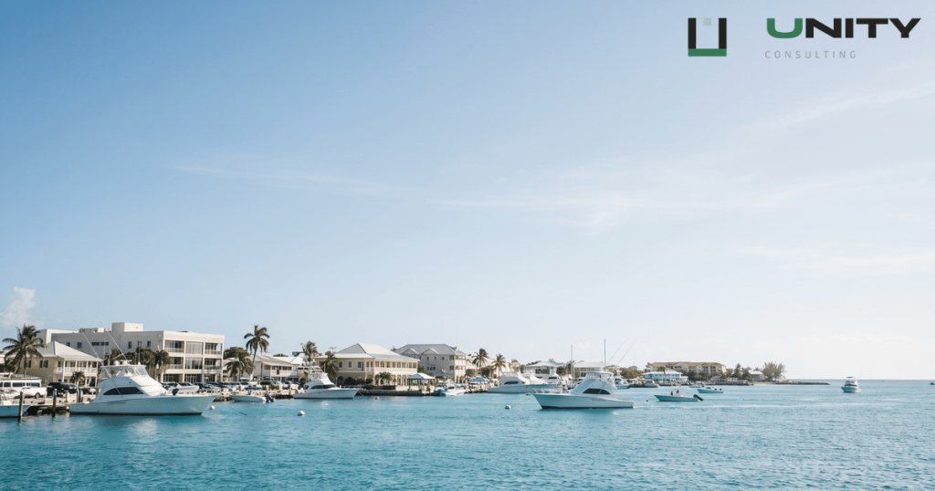 A wide shot of George Town harbour, Grand Cayman, featuring turquoise water, boats, and low-rise commercial buildings under a clear blue sky.