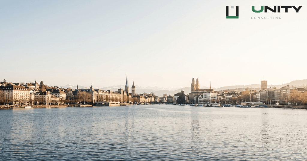 A wide-angle, deep focus view of the Zurich skyline across Lake Zurich with distant Alps under a clear sky.