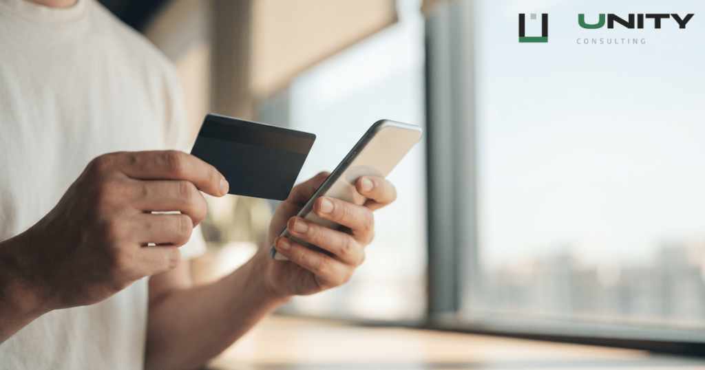 Hands holding a plain payment card and a smartphone in a brightly lit, blurred modern office environment.