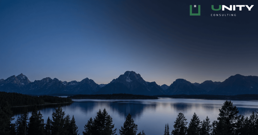 The Grand Teton mountain range at blue hour, silhouetted against a dark blue sky with a calm lake in the foreground.