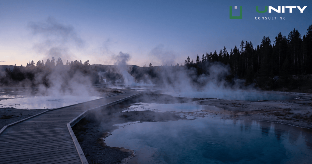 A wide view of a misty geyser basin in Yellowstone National Park at blue hour, with dark silhouetted pines.